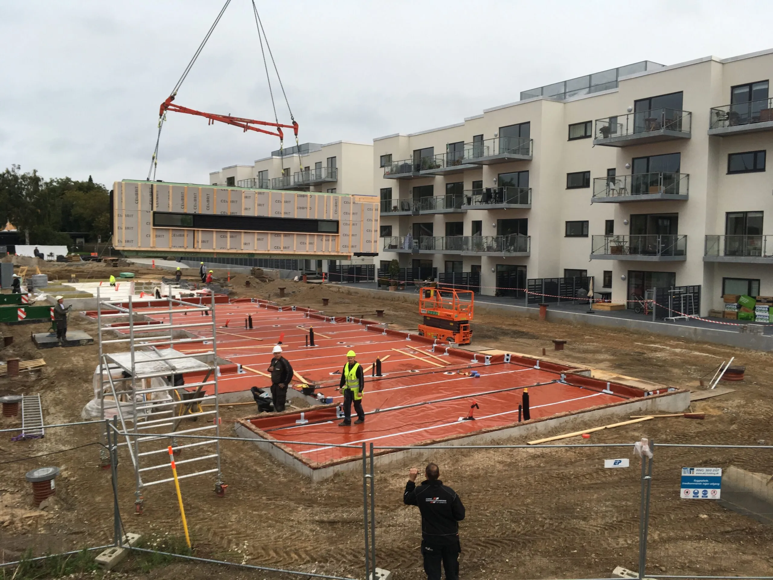 Byggeplads hvor kran løfter præfabrikeret modul over forberedt fundament med rødt underlag, færdig boligblok i baggrunden